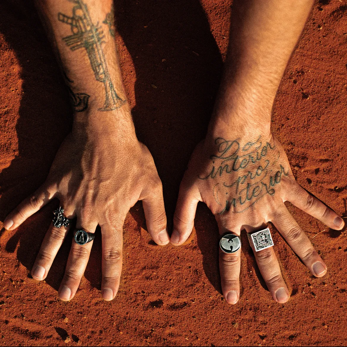 Mãos tatuadas com anéis apoiadas na terra vermelha